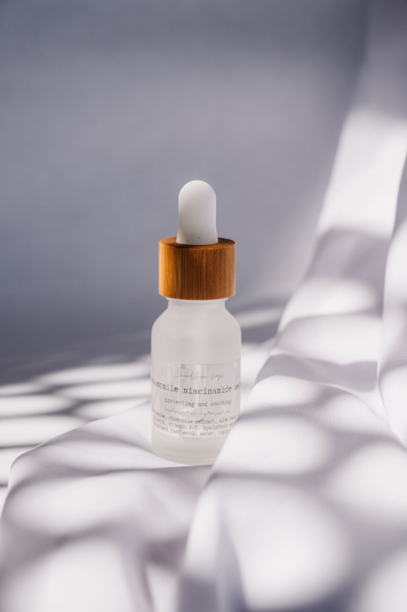 Może przedstawiać: Przezroczysta butelka ze szkła z bambusową nakrętką i kroplomierzem, zawierająca serum z etykietą "Gentle Niacinamide Serum, protecting and soothing". Butelka stoi na białej powierzchni z tkaniny.