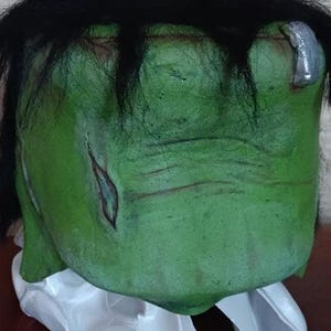 Puede incluir: Una máscara verde de Frankenstein con pelo negro y un perno plateado en el lateral de la cabeza. La máscara tiene una superficie texturizada y una base blanca.