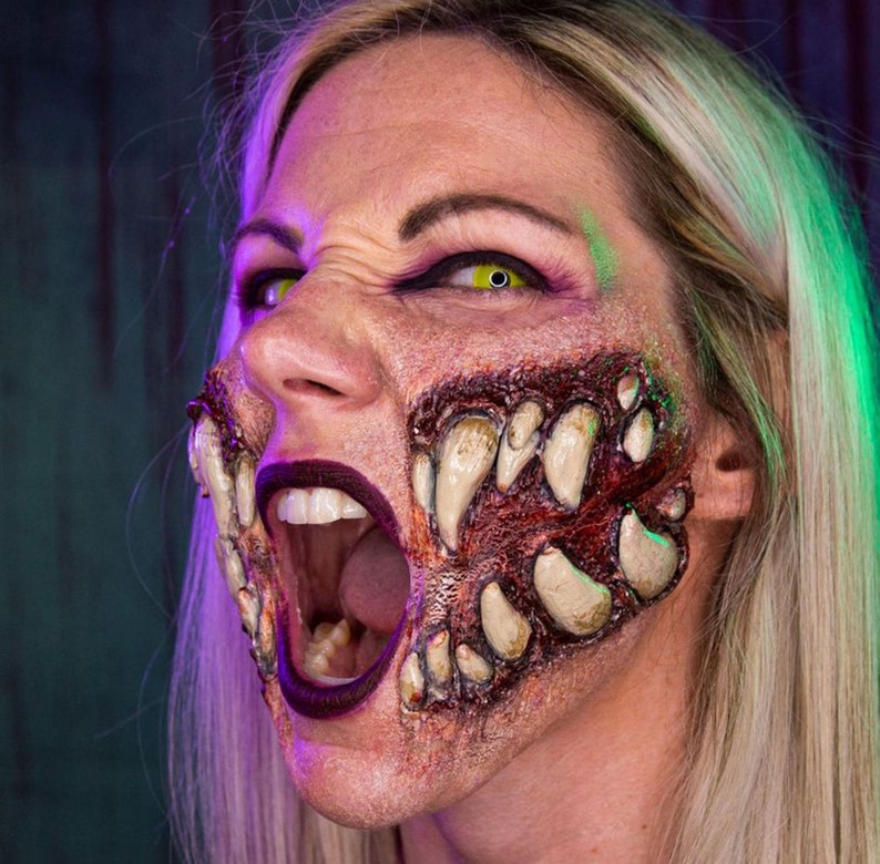 May include: A woman with blonde hair has a special effects makeup design on her face. The makeup is a realistic depiction of a monster's mouth with sharp teeth and a bloody, raw appearance.