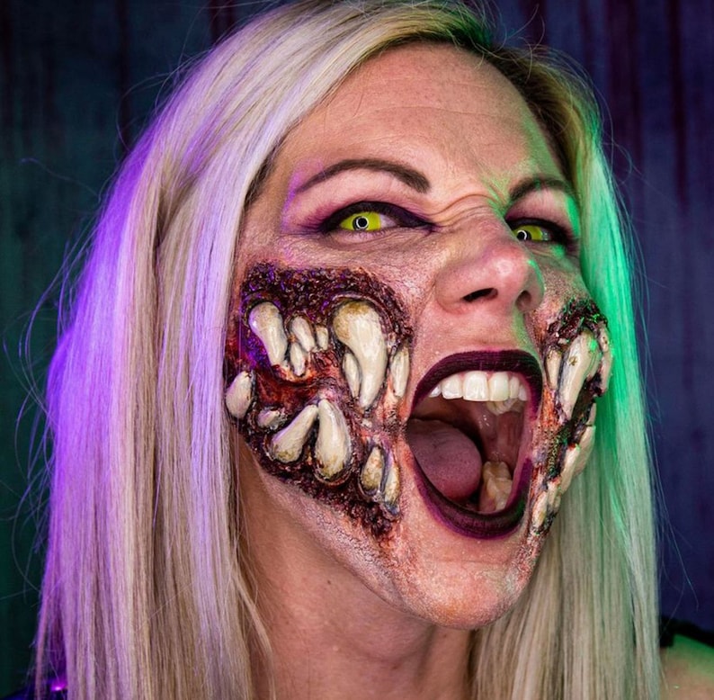 May include: A woman with blonde hair is wearing a special effects makeup design that makes her look like a monster. The makeup includes a large, open mouth with sharp teeth and a bloody, fleshy texture.