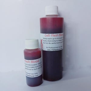 Może przedstawiać: Dwie butelki sztucznej krwi, jedna mała i jedna duża. Mniejsza butelka jest oznaczona etykietą "Flesh Blood", a większa butelka "Gell- Flesh Blood". Obie butelki są czerwone i mają białe nakrętki.