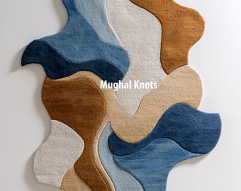 Puede incluir: Alfombra abstracta con una forma orgánica única. La alfombra presenta una mezcla de colores, incluyendo tonos de azul, marrón, beige y blanco roto. El texto "Mughal Knots" se muestra en la alfombra.