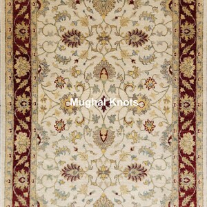 Puede incluir: Una alfombra de color crema con un borde rojo y intrincados patrones florales. La alfombra tiene un diseño tradicional y presenta un motivo central con el texto "Mughal Knots".