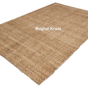 K&ouml;nnte beinhalten: Ein Naturfaser-Teppich aus Jute, einem nachhaltigen und strapazierf&auml;higen Material. Der Teppich hat eine gewebte Textur und eine hellbraune Farbe. Der Text "Mughal Knots" ist in Wei&szlig; auf den Teppich gedruckt.