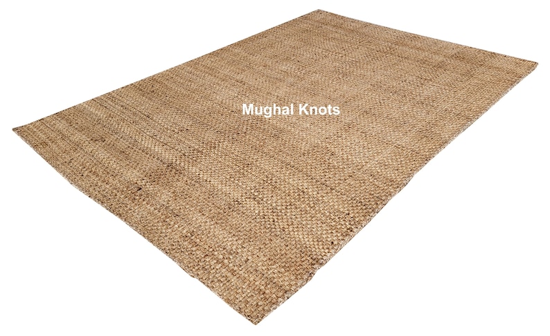K&ouml;nnte beinhalten: Ein Naturfaser-Teppich aus Jute, einem nachhaltigen und strapazierf&auml;higen Material. Der Teppich hat eine gewebte Textur und eine hellbraune Farbe. Der Text "Mughal Knots" ist in Wei&szlig; auf den Teppich gedruckt.