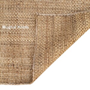 K&ouml;nnte beinhalten: Nahaufnahme eines Naturfaser-Teppichs mit gewebter Textur. Der Teppich ist umgeklappt, wodurch die Unterseite sichtbar wird. Der Text "Mughal Knots" ist auf dem Teppich zu sehen.