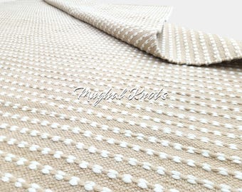 May include: Beige and white woven rug with a dotted pattern. The rug is folded over, revealing the underside. The text "Mughal Knots" is visible in the centre of the rug.