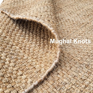 K&ouml;nnte beinhalten: Nahaufnahme eines Naturfaser-Teppichs mit gewebter Textur. Der Teppich ist hellbraun und hat einen leicht ausgefransten Rand. Der Text "Mughal Knots" ist in der unteren rechten Ecke des Bildes sichtbar.