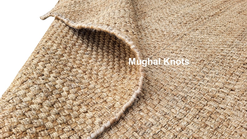 K&ouml;nnte beinhalten: Nahaufnahme eines Naturfaser-Teppichs mit gewebter Textur. Der Teppich ist hellbraun und hat einen leicht ausgefransten Rand. Der Text "Mughal Knots" ist in der unteren rechten Ecke des Bildes sichtbar.