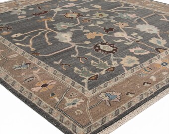 May include: A square area rug with a floral pattern. The rug features a gray center with a repeating floral design in shades of brown, beige, and blue. The border is a contrasting brown with a repeating floral pattern. The rug has a fringe edge.