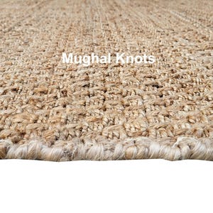 K&ouml;nnte beinhalten: Nahaufnahme eines Naturfaser-Teppichs mit gewebter Textur. Der Teppich hat eine hellbraune Farbe und einen ausgefransten Rand. Der Text "Mughal Knots" ist im Bild sichtbar.