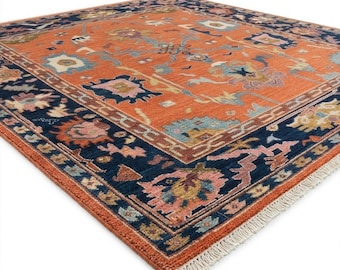 May include: An orange and navy blue area rug with a detailed floral pattern. The rug has a navy blue border with a repeating floral design and a fringe on one edge. The center of the rug is orange with a variety of floral shapes in pink, blue, and yellow.
