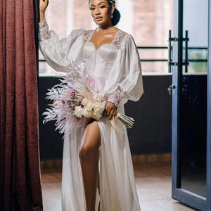 Puede incluir: Una novia con un vestido de novia de satén blanco con un corpiño de encaje y mangas largas. El vestido tiene una abertura en la pierna hasta el muslo y la novia lleva un ramo de flores rosas y blancas.