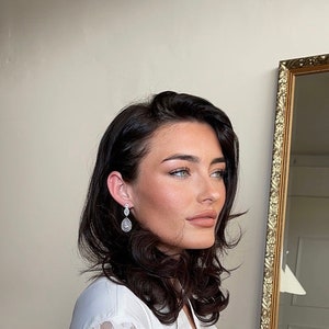 May include: A woman with dark brown hair wearing a white shirt and dangling diamond earrings. She is looking to the side and has her hair styled in loose waves.