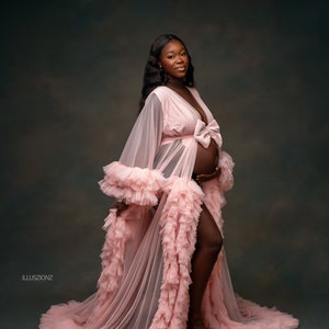May include: A woman in a light pink maternity robe with a plunging neckline and a bow at the waist. The robe has ruffled detailing on the sleeves and hem. The background is a muted grey. The robe's train trails on the floor.