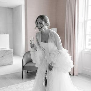 Może przedstawiać: Biała suknia ślubna z falbanami na rękawach i pasującą sukienką. Szlafrok jest otwarty z przodu, odsłaniając sukienkę pod spodem. Kobieta trzyma kieliszek szampana i się śmieje. Pokój ma jasnoróżowe ściany i neutralny dywan.