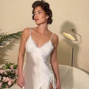 May include: White satin slip dress with lace trim on the neckline and hem. The dress features spaghetti straps and a side slit with tie detailing. The model is wearing the dress in a room with a neutral background.