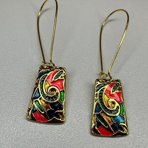 May include: A pair of gold-coloured drop earrings. Each earring has a rectangular pendant with a colourful, abstract design in red, green, blue, and yellow. The earrings have a hook closure.
