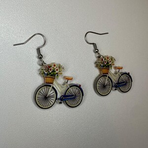 May include: A pair of bicycle earrings. Each earring depicts a white bicycle with a basket of flowers. The bicycle has black wheels, blue mudguards, and a brown basket. The earrings have silver-coloured fish hook ear wires.