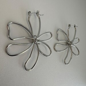 May include: A pair of silver flower-shaped hoop earrings. Each earring has an openwork design with five petals. The earrings are made of a shiny, metallic material. They are displayed against a light background.