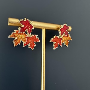 May include: A pair of gold-tone earrings featuring clusters of maple leaves in shades of red, orange, and yellow enamel. Each earring consists of three distinct leaf shapes with metallic gold outlines, displayed on a gold-coloured T-bar stand.