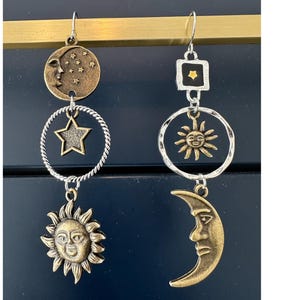 以下が含まれることがあります： 銀と金の天体モチーフのイヤリングのペア。片方のイヤリングは三日月、星、顔のある太陽。もう片方のイヤリングは星、顔のある太陽、三日月のある正方形です。