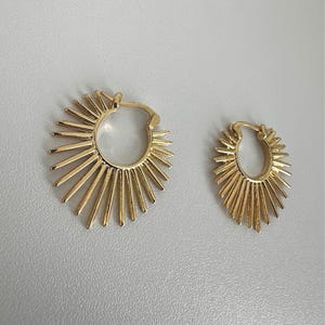 Peut inclure: Une paire de boucles d'oreilles créoles dorées avec un motif en forme de soleil composé de pointes rayonnantes s'étendant depuis la base circulaire. Les boucles d'oreilles possèdent un mécanisme de fermeture à charnière et une finition métallique polie.