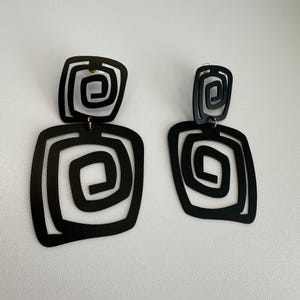 May include: Black geometric earrings featuring a square shape and a spiral design. The earrings have a post closure and a gold-coloured connecting piece. A bold statement piece.