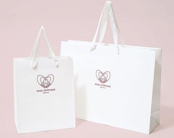 May include: Two white paper shopping bags with white ribbon handles. Each bag features a dark brown logo of a baby inside a heart formed by two hands, with the text ANGEL PATRONAGE zakka shop printed in dark brown lettering below the logo.