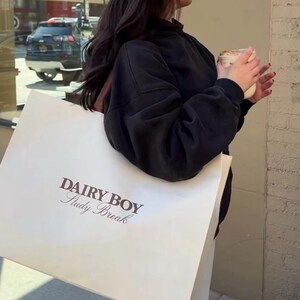 May include: A person holding a white tote bag with the text "Dairy Boy Study Break" printed on it. The person is wearing a black sweatshirt, blue jeans, and a white baseball cap with a green brim.