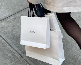 May include: White shopping bags with black handles are held by a person. The bags have the word "REFY" printed on them. The person is wearing a white jacket and black tights.