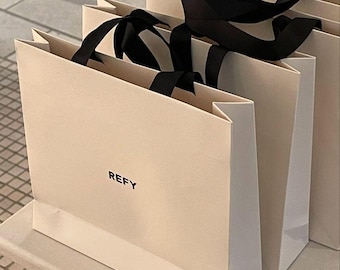 May include: Stack of three beige shopping bags with black handles and black ribbon bows. The bottom bag has the word "REFY" printed in black on the front.