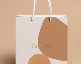 May include: A rectangular paper gift bag with white rope handles. The bag features a cream-colored background with large, abstract tan and light brown shapes printed on the front. The word Breeze is printed in a gold serif font in the center.