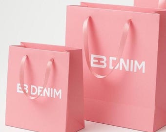 May include: Pink shopping bags with white text that reads "EB DENIM" and "BNIM". The bags have matching pink ribbon handles. The bags are in different sizes, with the smaller bag in the foreground.