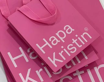 May include: Three pink shopping bags stacked on top of each other. The bags have a white logo that says "Hapa Kristin" with a small circle and a letter "K" inside the circle.