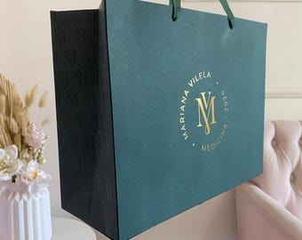 May include: A teal shopping bag with a gold logo that reads "Mariana Vilela Medicina 2024". The bag has a black textured side and green ribbon handles. The bag is sitting on a white surface.