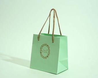 May include: A light green paper shopping bag with a gold-colored logo and rope handles. The logo features a circular wreath design with text. The bag is empty and stands on a light green surface.