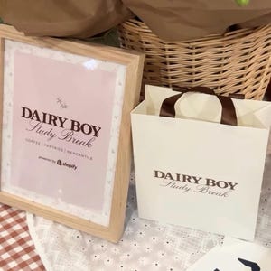 May include: A white paper bag with brown handles and the text "DAIRY BOY Study Break" printed on it. The bag is sitting on a white tablecloth with a floral pattern.