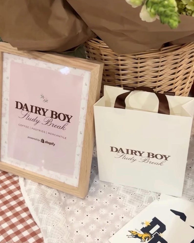 May include: A white paper bag with brown handles and the text "DAIRY BOY Study Break" printed on it. The bag is sitting on a white tablecloth with a floral pattern.