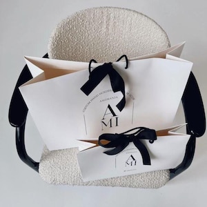 May include: Two white paper shopping bags with black ribbon handles and the text "AMI" printed on them. The bags are on a chair with a beige seat and black frame. The text "CUSTOM HAND HOLDERS" and "MOSCOW" are also printed on the bags.