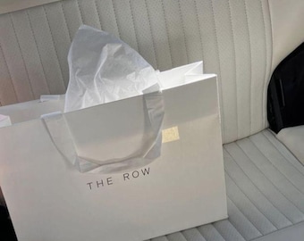 May include: A white shopping bag with the text "THE ROW" printed on the front. The bag has white handles and is filled with white tissue paper. The bag is sitting on a white, quilted car seat.