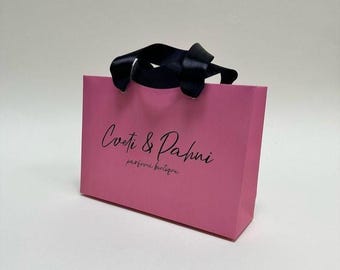 Puede incluir: Una bolsa de regalo rectangular de papel rosa con asas de cinta de satén negra. La parte frontal de la bolsa muestra el texto impreso en negro Coeti & Pahni parfume boutique en una combinación de fuente cursiva y sans-serif centrada.