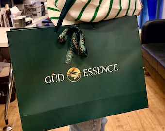 May include: A large, dark green shopping bag with the words "GÜD ESSENCE" in white. The bag has a gold logo and a green ribbon tied in a bow. The bag is being held by someone wearing a striped sweater.