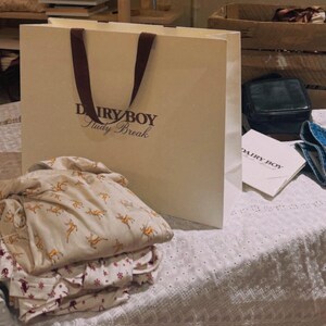 May include: A white paper shopping bag with brown handles and the text "Dairy Boy Study Break" printed on the front. The bag is sitting on a white lace tablecloth with a stack of white and brown patterned shirts.