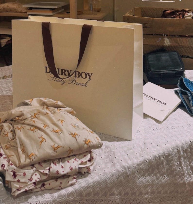May include: A white paper shopping bag with brown handles and the text "Dairy Boy Study Break" printed on the front. The bag is sitting on a white lace tablecloth with a stack of white and brown patterned shirts.