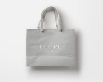Puede incluir: Una bolsa de papel de color gris claro con dos asas de cinta a juego. La parte frontal de la bolsa presenta el texto blanco LEÓRE centrado sobre el texto blanco más pequeño HOME COLLECTION en una fuente serif limpia y elegante.