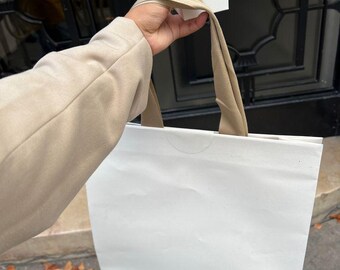 May include: A white shopping bag with the text "JACQUEMUS" printed on the front. A smaller white box with the same text is being held in the hand of a person.