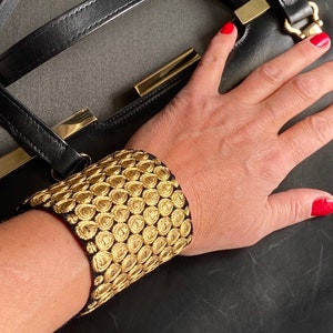 Pode incluir: Uma bolsa de mão de couro preto com fecho dourado e um bracelete dourado com padrão dourado e preto.