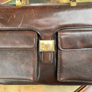 May include: A brown leather briefcase with gold hardware and multiple pockets. The briefcase has a worn look and is closed.