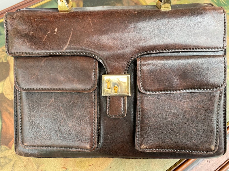 May include: A brown leather briefcase with gold hardware and multiple pockets. The briefcase has a worn look and is closed.